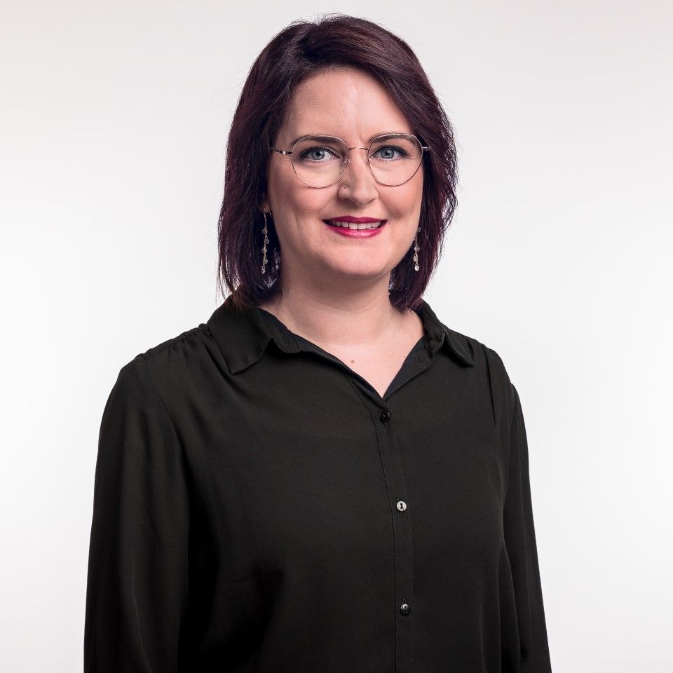 Eine Frau mit langen braunen Haaren und schwarzem Oberteil lächelt vor weißem Hintergrund.