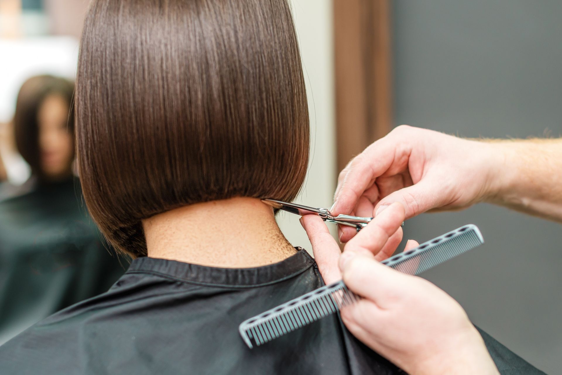 Eine Friseurin schneidet mit Schere und Kamm die hinteren Haare eines dunkelbraunen Bob-Haarschnitts.