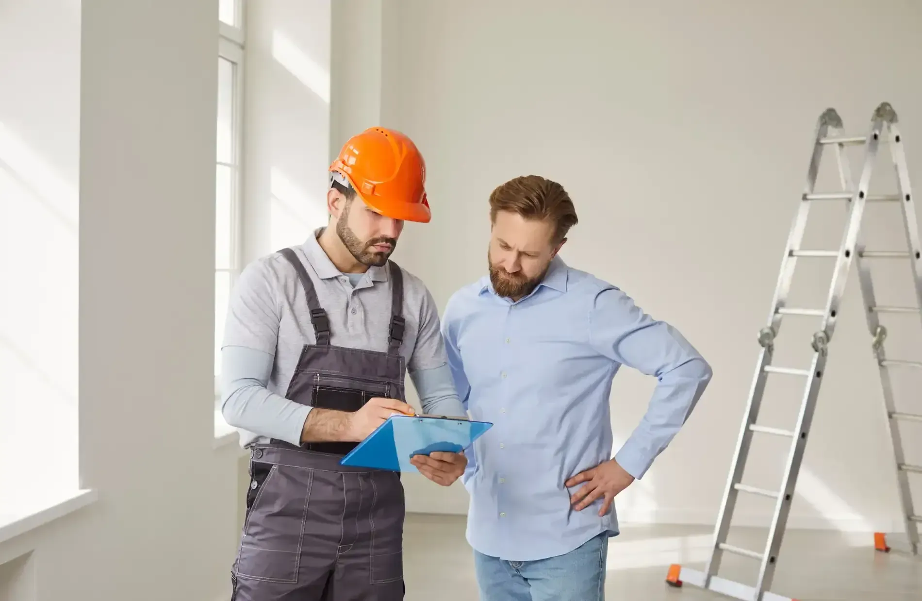 Un obrero de la construcción con casco naranja muestra una carpeta a un hombre con camisa azul; al fondo se ve una escalera.