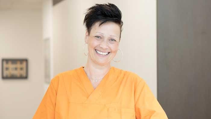 Eine Frau in einem orangefarbenen OP-Kittel lächelt in die Kamera.