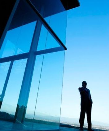 La silueta de una persona con traje se alza junto a una gran pared de cristal, con un cielo azul brillante de fondo.