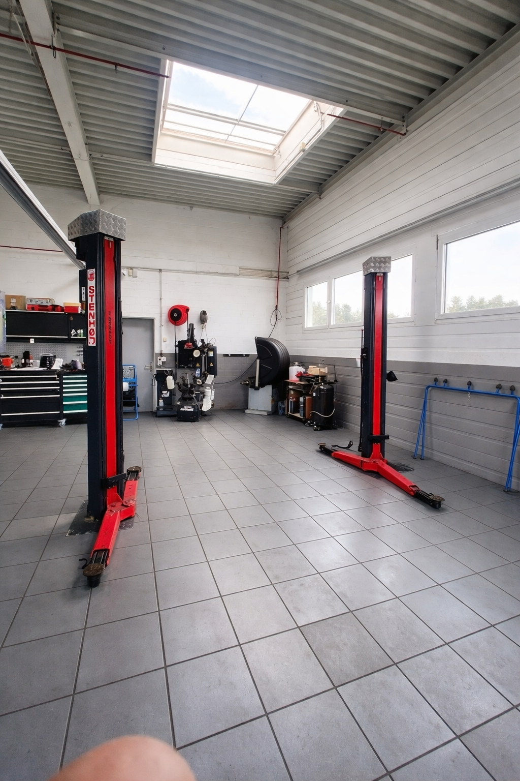 Eine Autowerkstatt mit zwei roten Hebebühnen, grauen Fliesenböden, weissen Wänden und einem grossen Dachfenster.