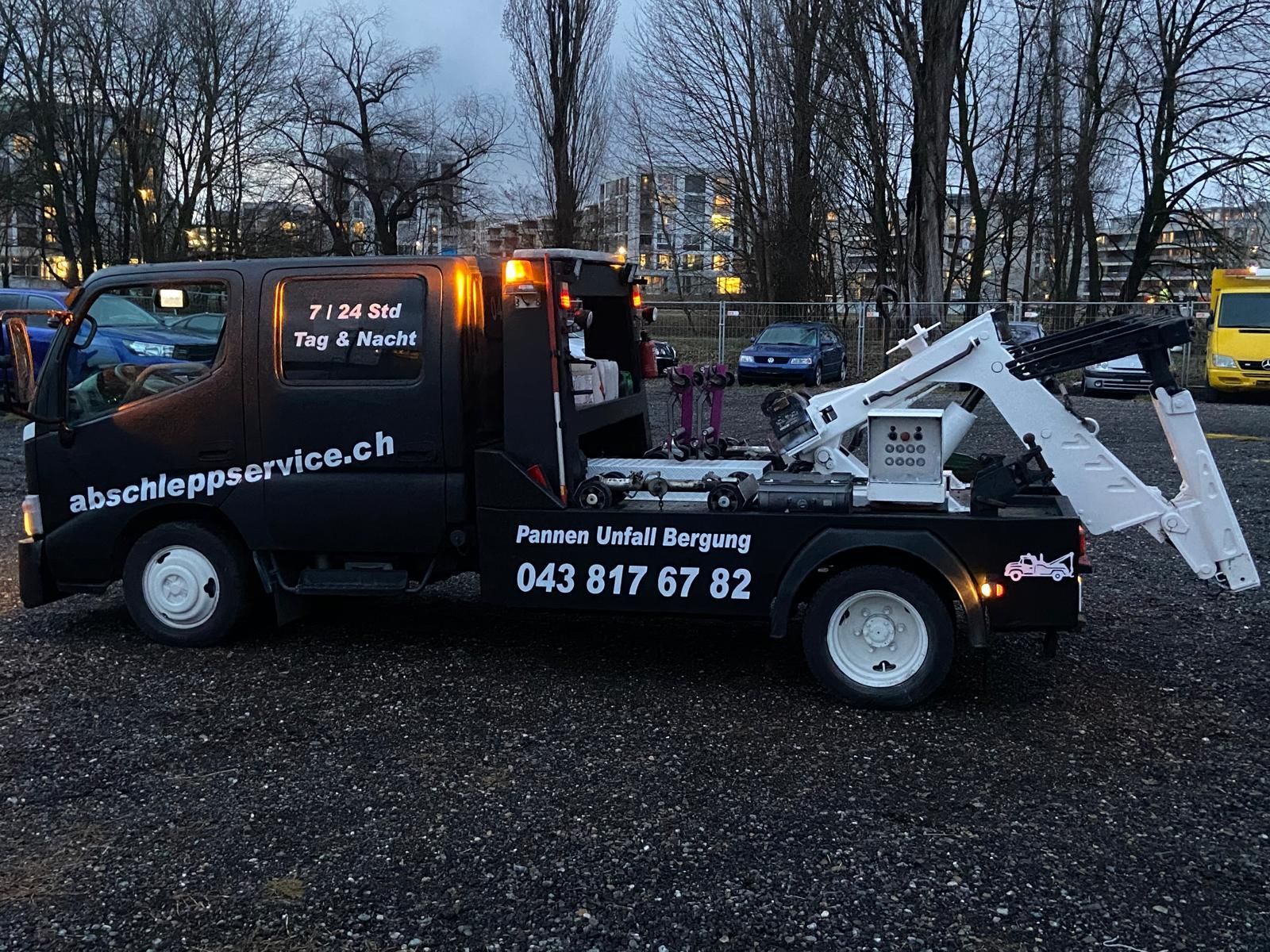 Ein Arbeiter in Warnweste und blauer Arbeitshose befestigt einen Autoreifen mit einem roten Gurt auf der Ladefläche eines Abschleppwagens.