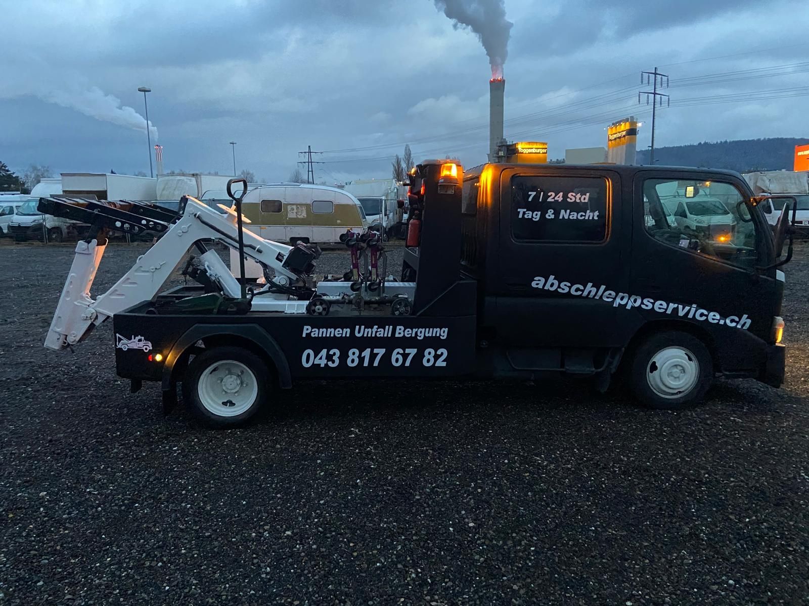 Ein Arbeiter in blauem Overall benutzt einen orangefarbenen Kranwagen, um einen schwarzen Geländewagen auf einen Tieflader in einer Stadtstrasse zu heben.