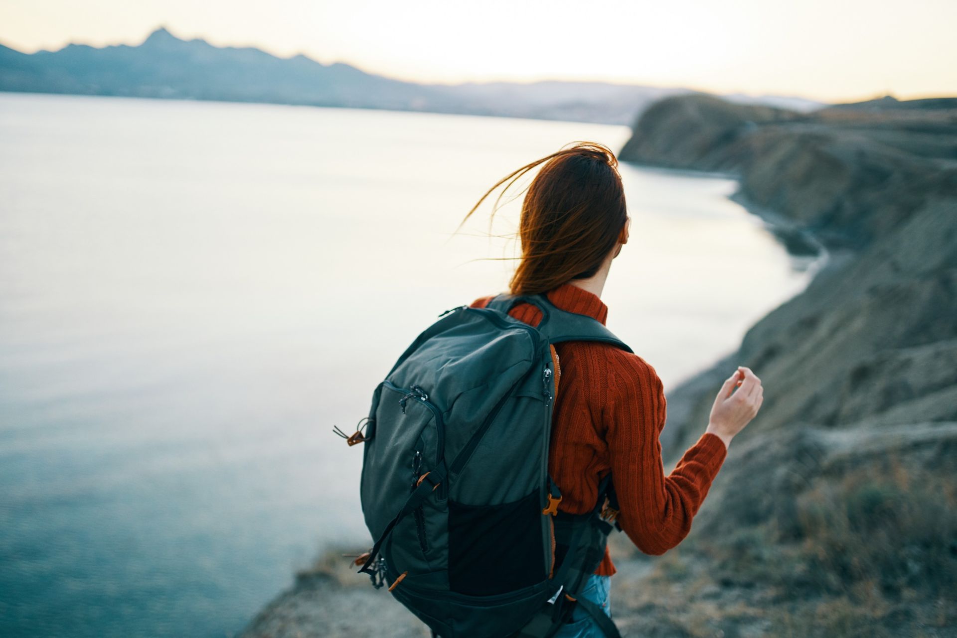 Una mujer con una mochila está parada en un acantilado con vista a un cuerpo de agua.