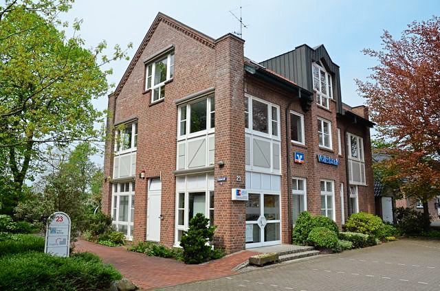 Ein großes Backsteingebäude mit vielen Fenstern ist von Bäumen und Büschen umgeben.