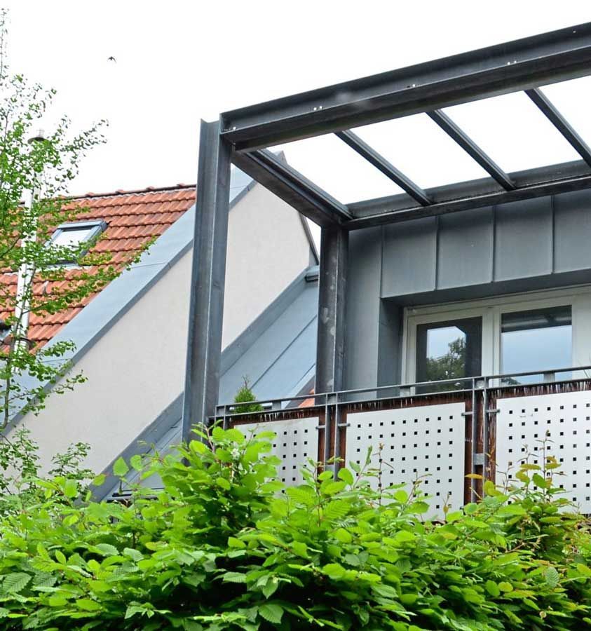 Ein Balkon mit Glasdach ist von grünen Büschen umgeben