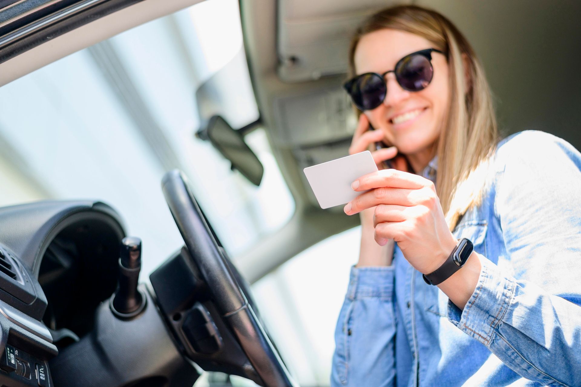 Mujer con gafas de sol sentada en un coche, sosteniendo una tarjeta de crédito y sonriendo.