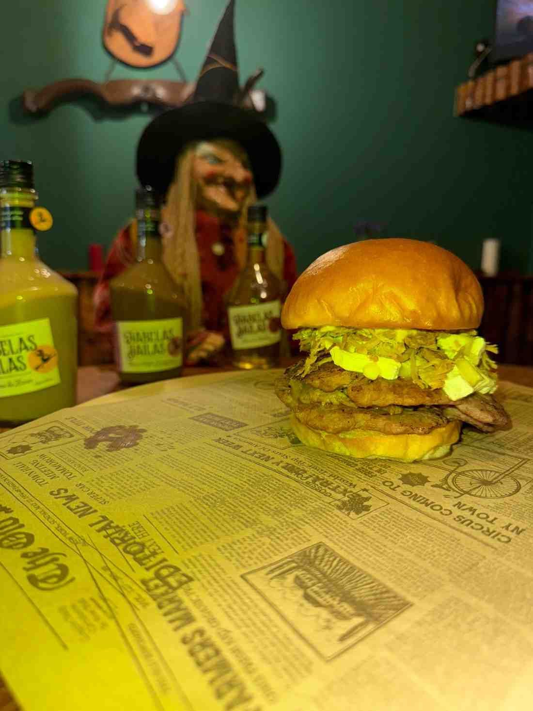 Hamburguesa sobre papel de periódico delante de decoración de brujas y botellas de líquido verde.