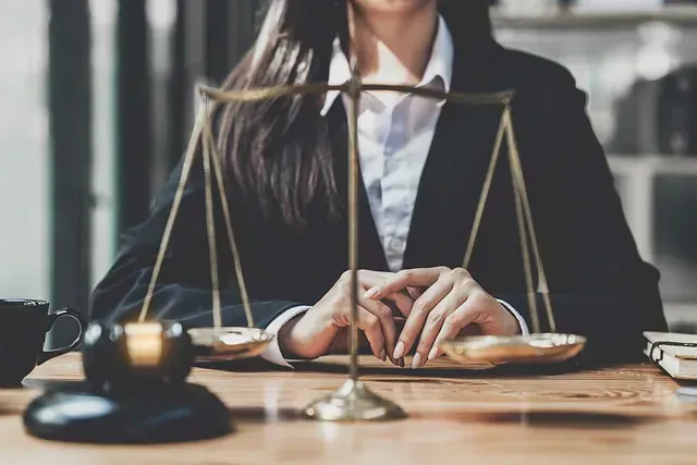 Una persona con un blazer se sienta detrás de un escritorio con una balanza de justicia y un mazo; el foco está en las manos y la balanza.