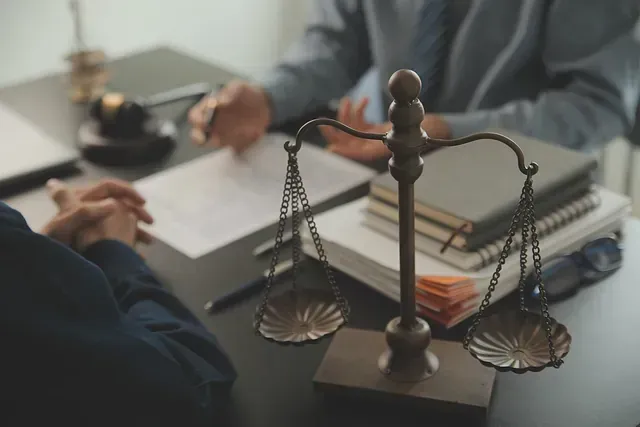 Una balanza de justicia en primer plano con personas sentadas en un escritorio, documentos y libros de leyes.