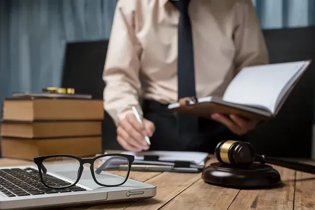 Persona con vestimenta formal escribiendo en un cuaderno, mazo y libros sobre un escritorio, computadora portátil y gafas en primer plano.