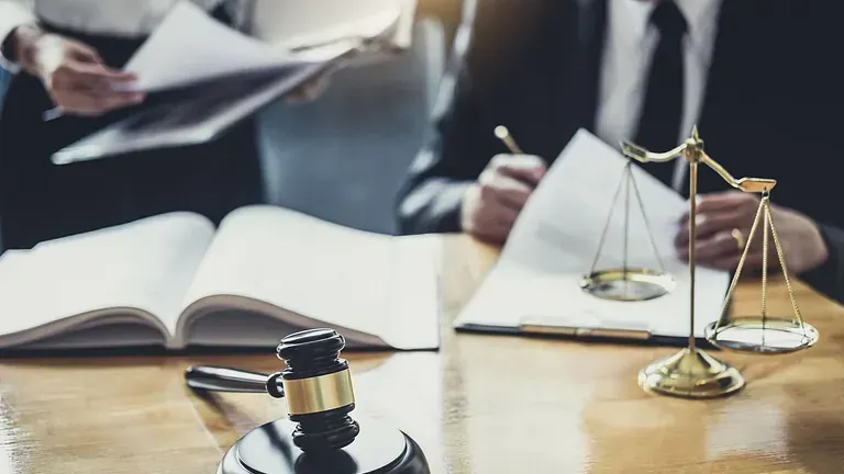 Dos abogados de traje revisan documentos con un mazo y una balanza de la justicia sobre un escritorio en primer plano.