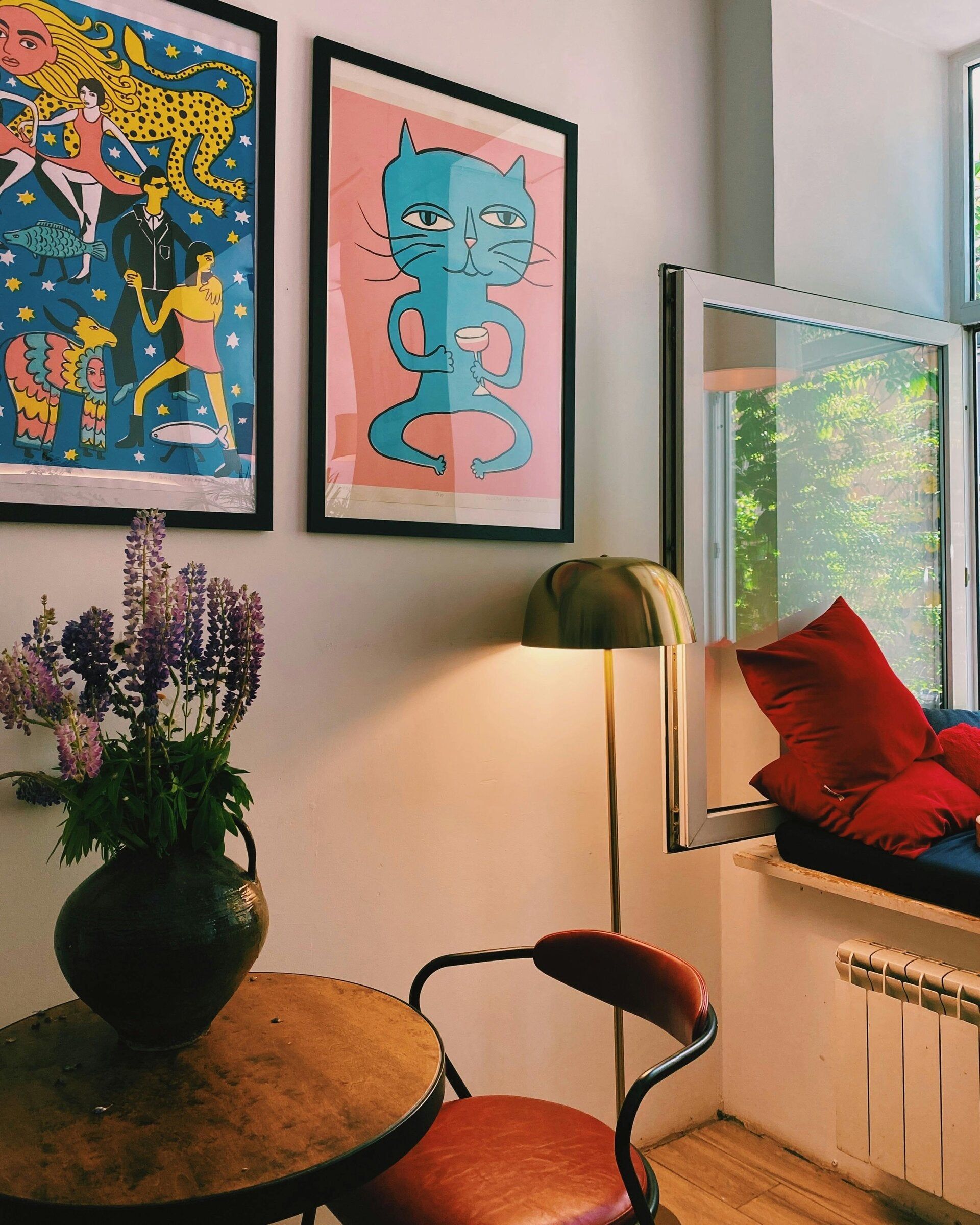 Un coin douillet avec une table ronde en bois, un vase de fleurs, une chaise rouge, un lampadaire doré et deux estampes encadrées
