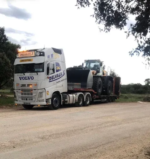 Un camion Volvo transporte un gros engin de chantier sur une remorque à plateau, sur un chemin de terre.