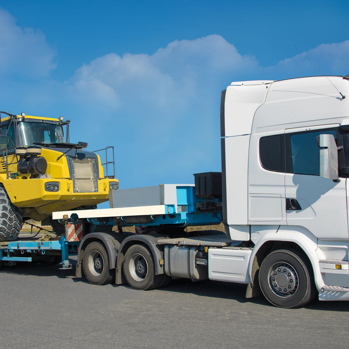 Un semi-remorque blanc transporte du matériel de construction jaune sur une remorque plateau bleue, sous un ciel bleu.