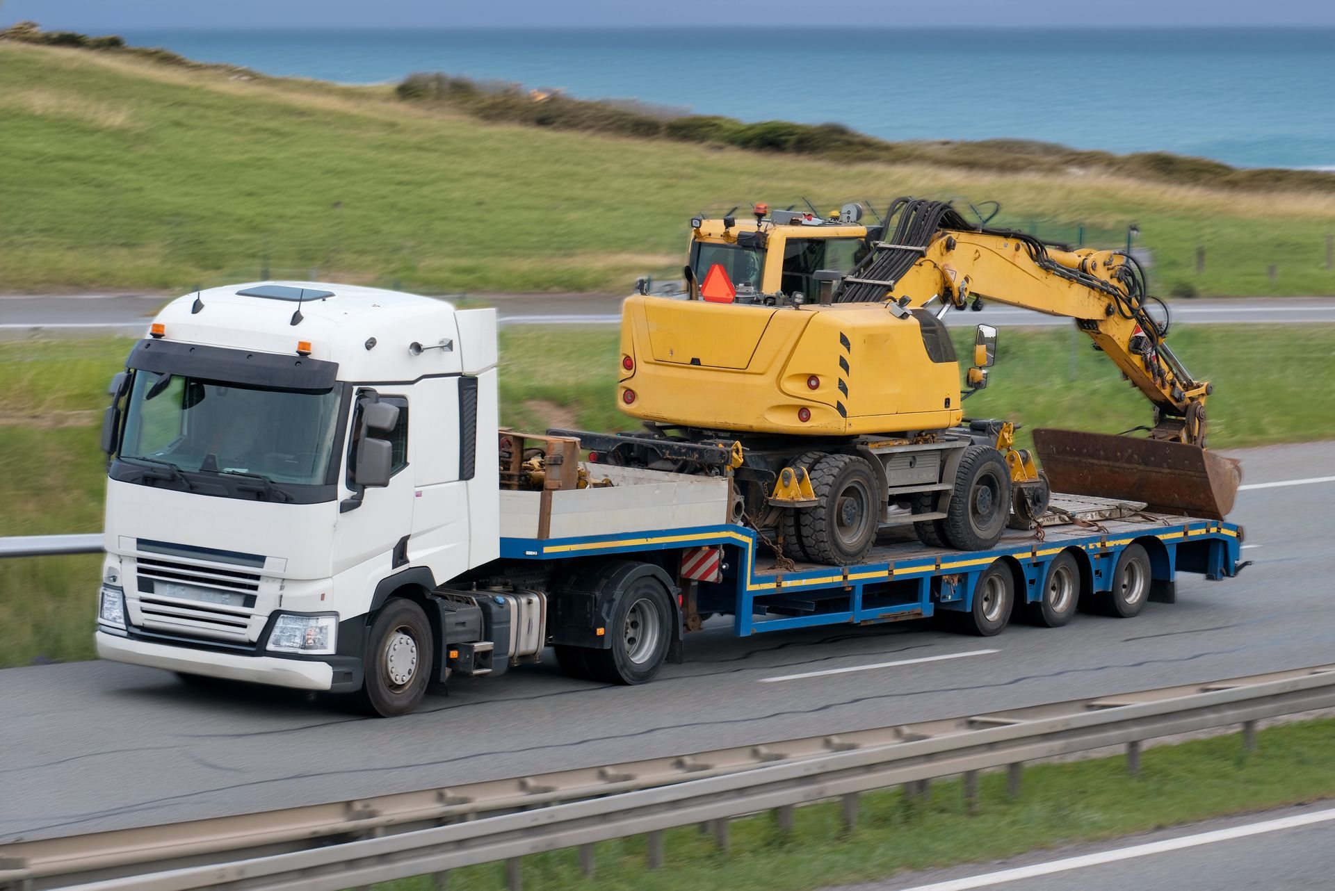 Un semi-remorque blanc transporte une excavatrice jaune sur une remorque à plateau, sur une autoroute.