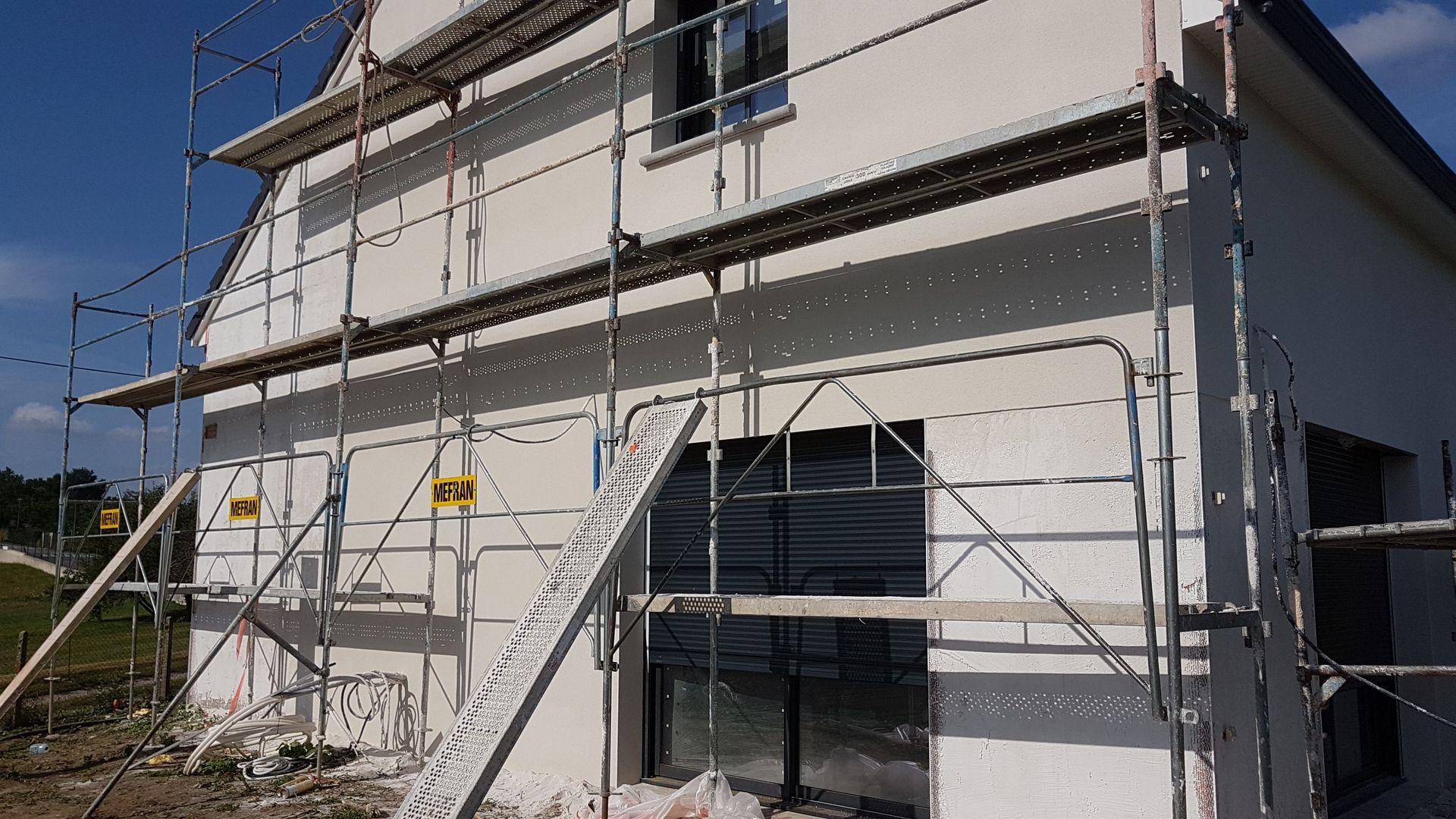 Échafaudages de chantier sur une maison à deux étages aux murs en stuc blanc, sous un ciel bleu.