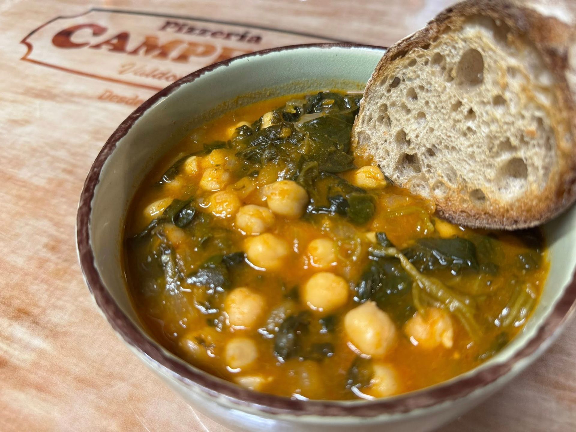 Sopa de garbanzos y col rizada en un recipiente de cerámica con una rebanada de pan sobre una superficie de madera.