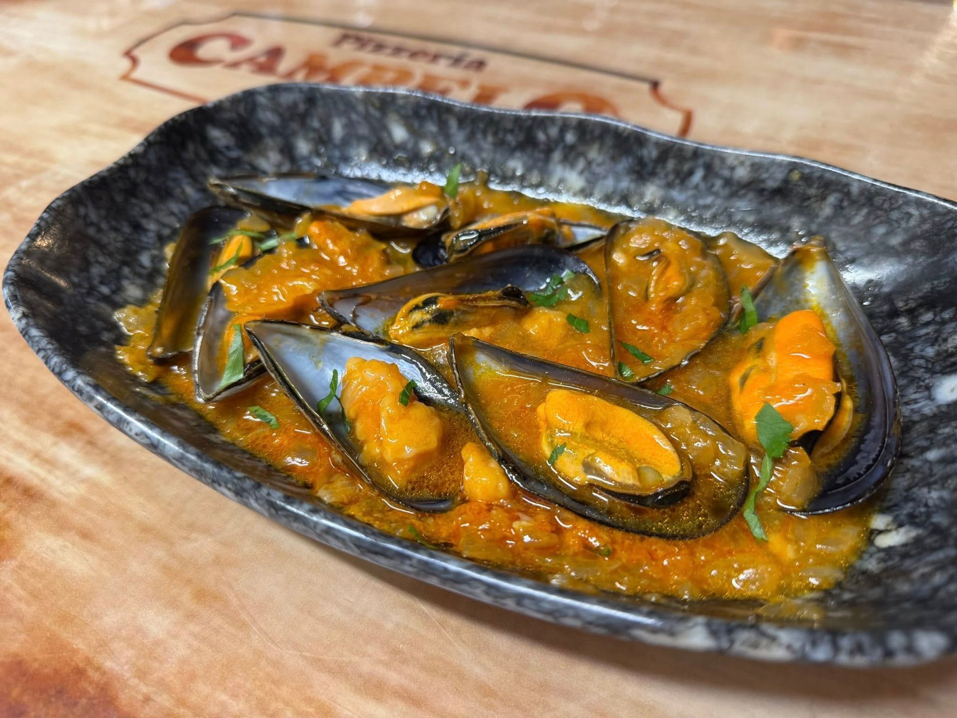 Mejillones en salsa de naranja sobre un plato oscuro con dibujos, colocado sobre una superficie de madera.