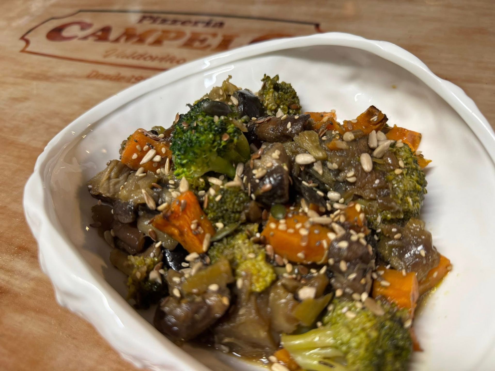 Salteado de verduras con brócoli, zanahorias y semillas de sésamo en un recipiente blanco, sobre una superficie de madera.