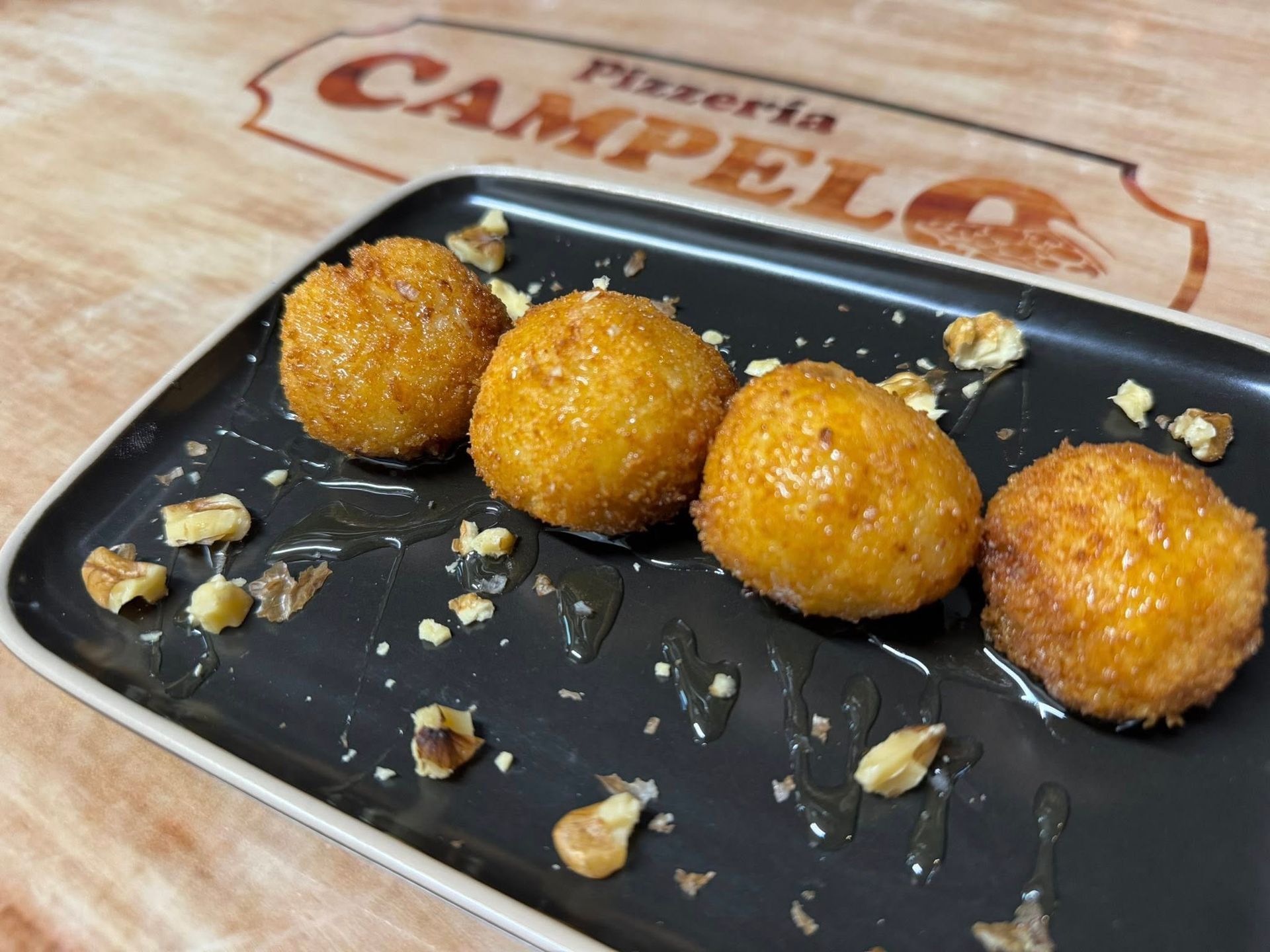 Cuatro bolas de queso fritas doradas rociadas con miel y trozos de nueces sobre un plato negro.