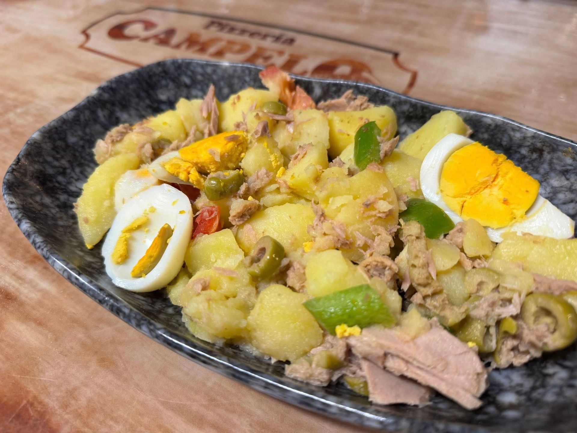 Ensalada de patata con atún, huevos y aceitunas en un plato gris. Superficie de madera, letrero del restaurante