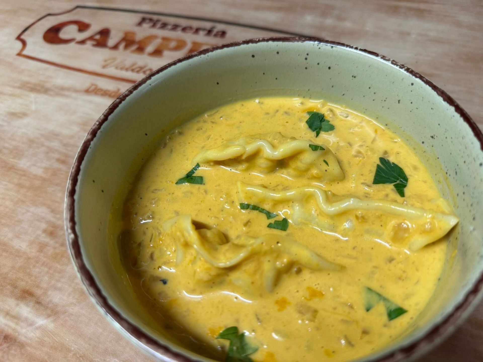 Sopa cremosa de color naranja con pasta y perejil en un recipiente rústico, con un cartel de pizzería al fondo.