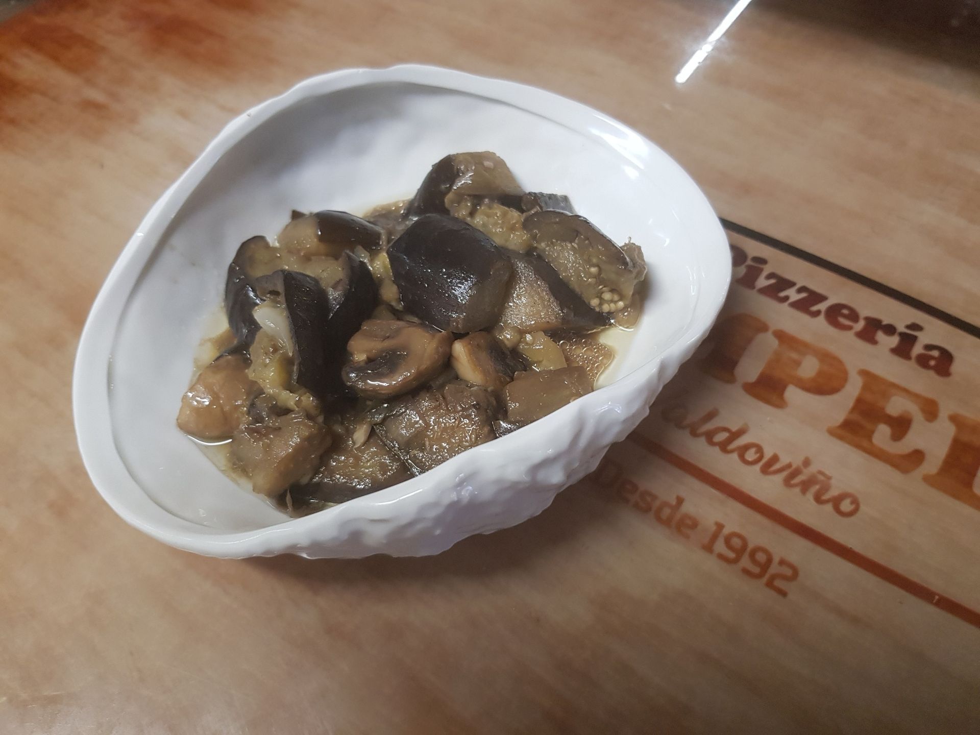 Plato de berenjenas y champiñones en un recipiente blanco de forma orgánica sobre una superficie de madera con el logotipo de un restaurante.