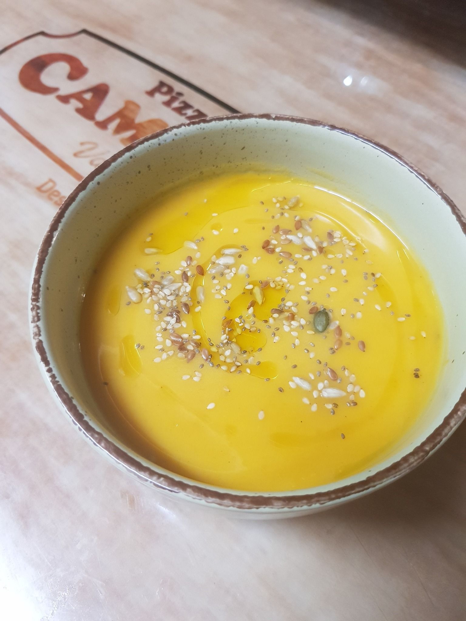 Un tazón de sopa cremosa de naranja con aceite de oliva y semillas, colocado sobre una mesa con una caja de pizza parcialmente visible.
