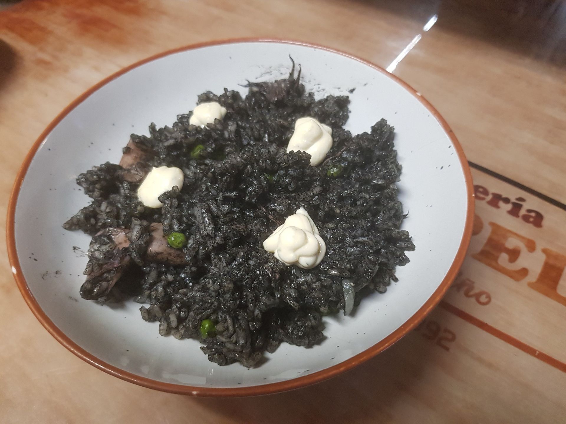 Plato de arroz negro cubierto con salsa blanca en un recipiente blanco, colocado sobre una superficie de madera.