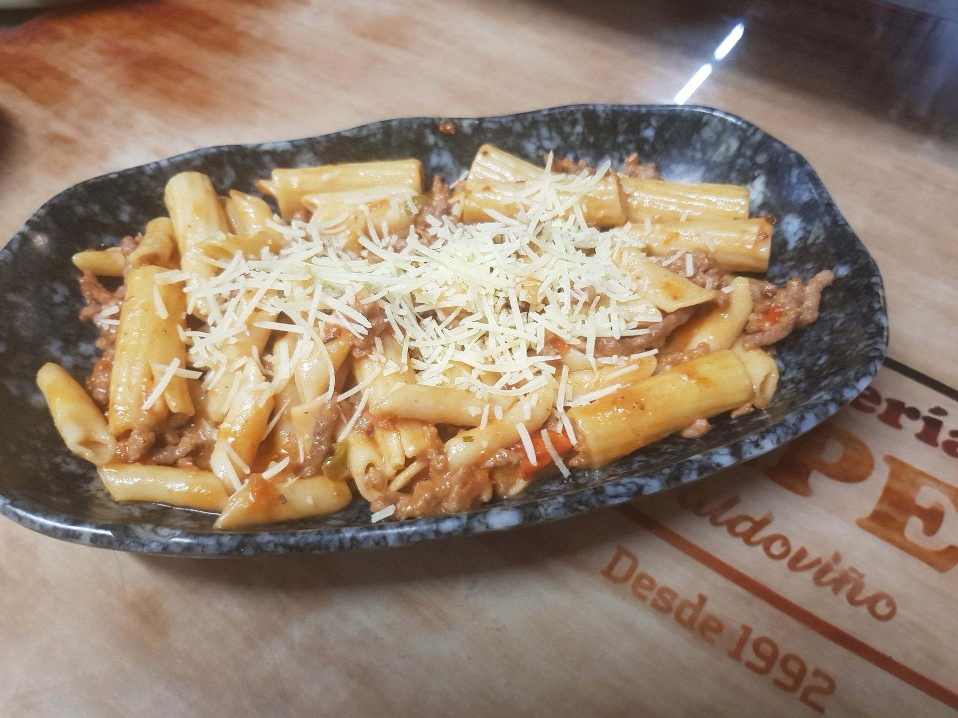 Plato de pasta con salsa de carne y queso rallado en un recipiente ovalado oscuro sobre una superficie de madera.