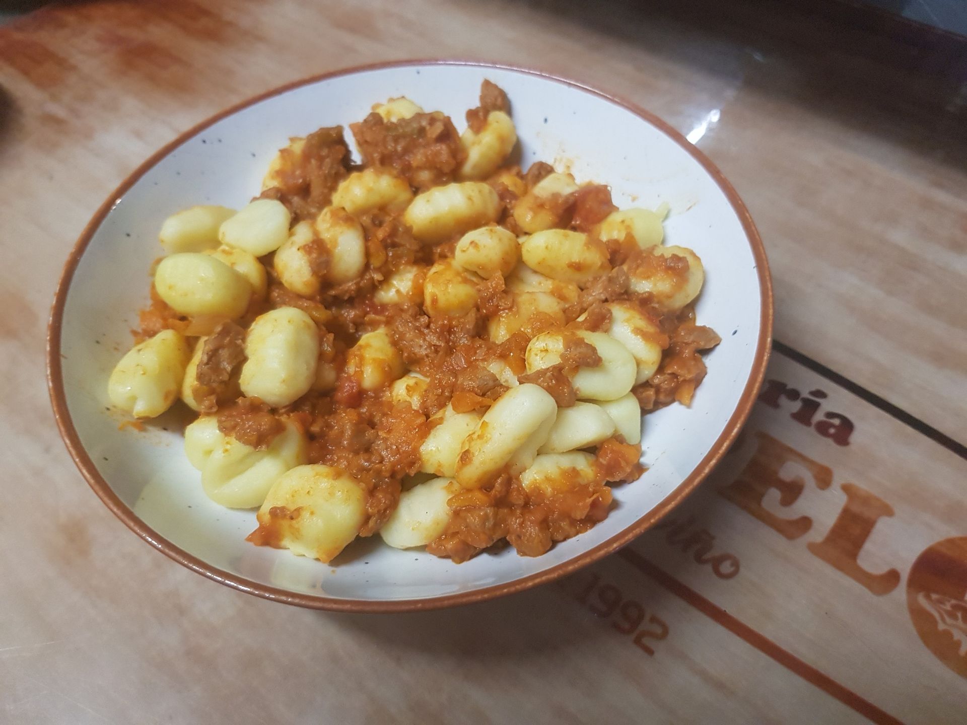 Pasta de ñoquis con salsa de carne en un recipiente blanco con borde marrón sobre una superficie de madera.