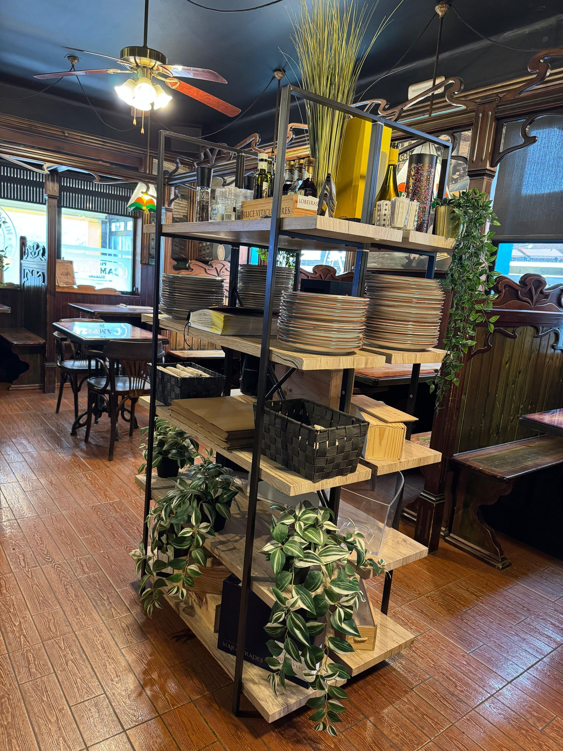 Estantería de un restaurante llena de platos, plantas y adornos. Suelo y mesas marrones.