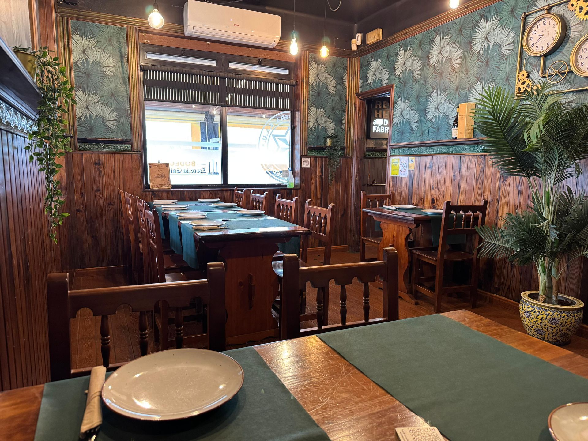 Interior del restaurante: mesas y sillas de madera, manteles verdes, papel pintado decorativo, plantas en macetas.