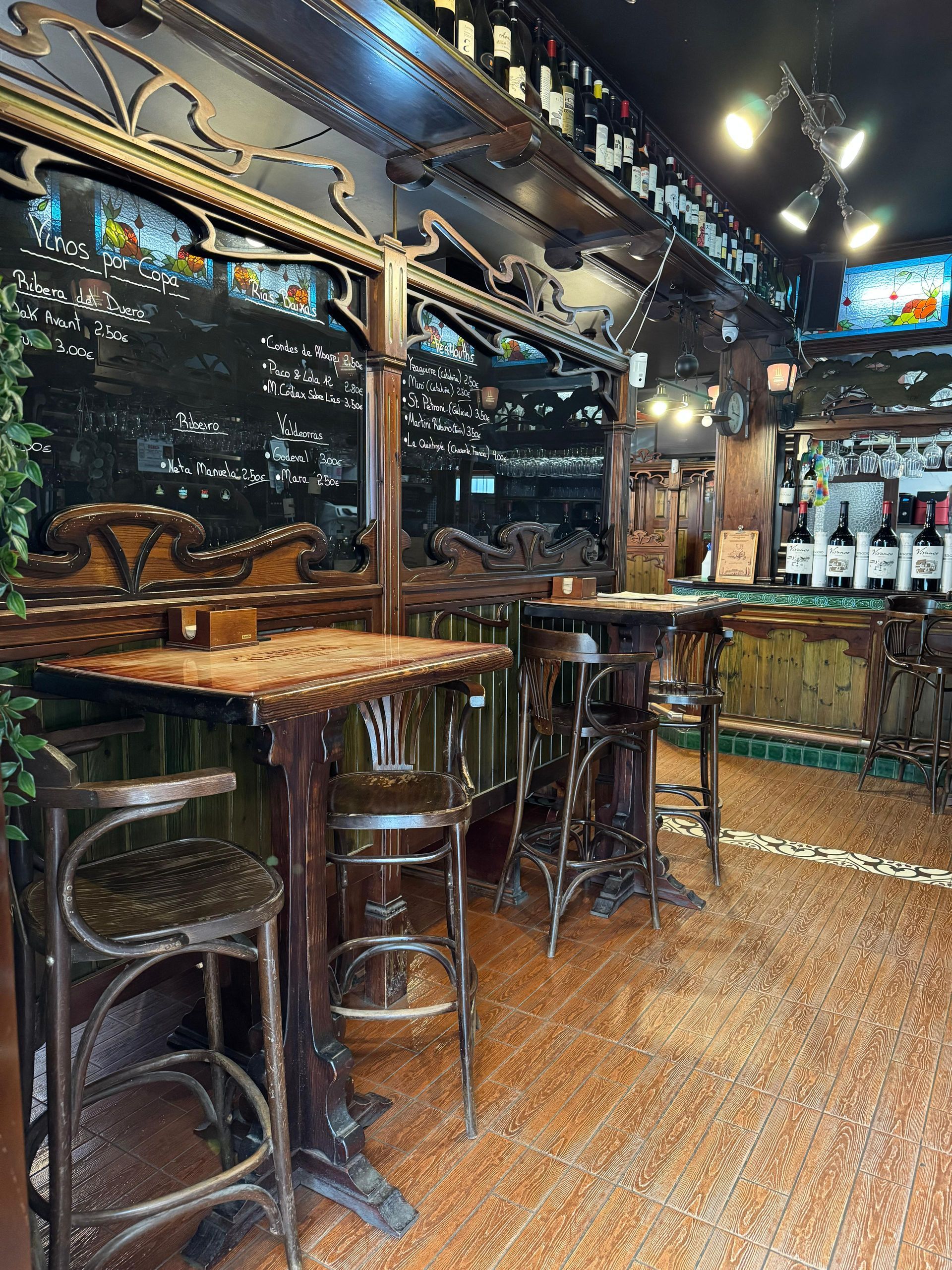 Interior de bar ornamentado con mesas de madera, taburetes y botellas de vino; iluminación tenue.