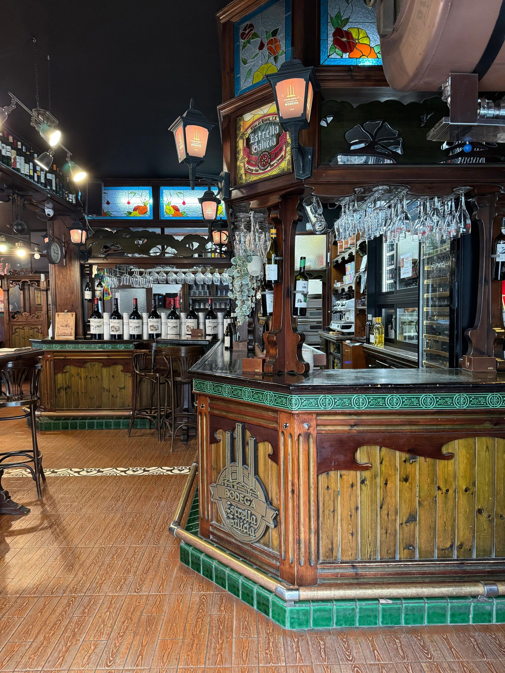 Interior de bar con mostradores de madera, estantes de botellas, lámparas de techo y suelo de baldosas.