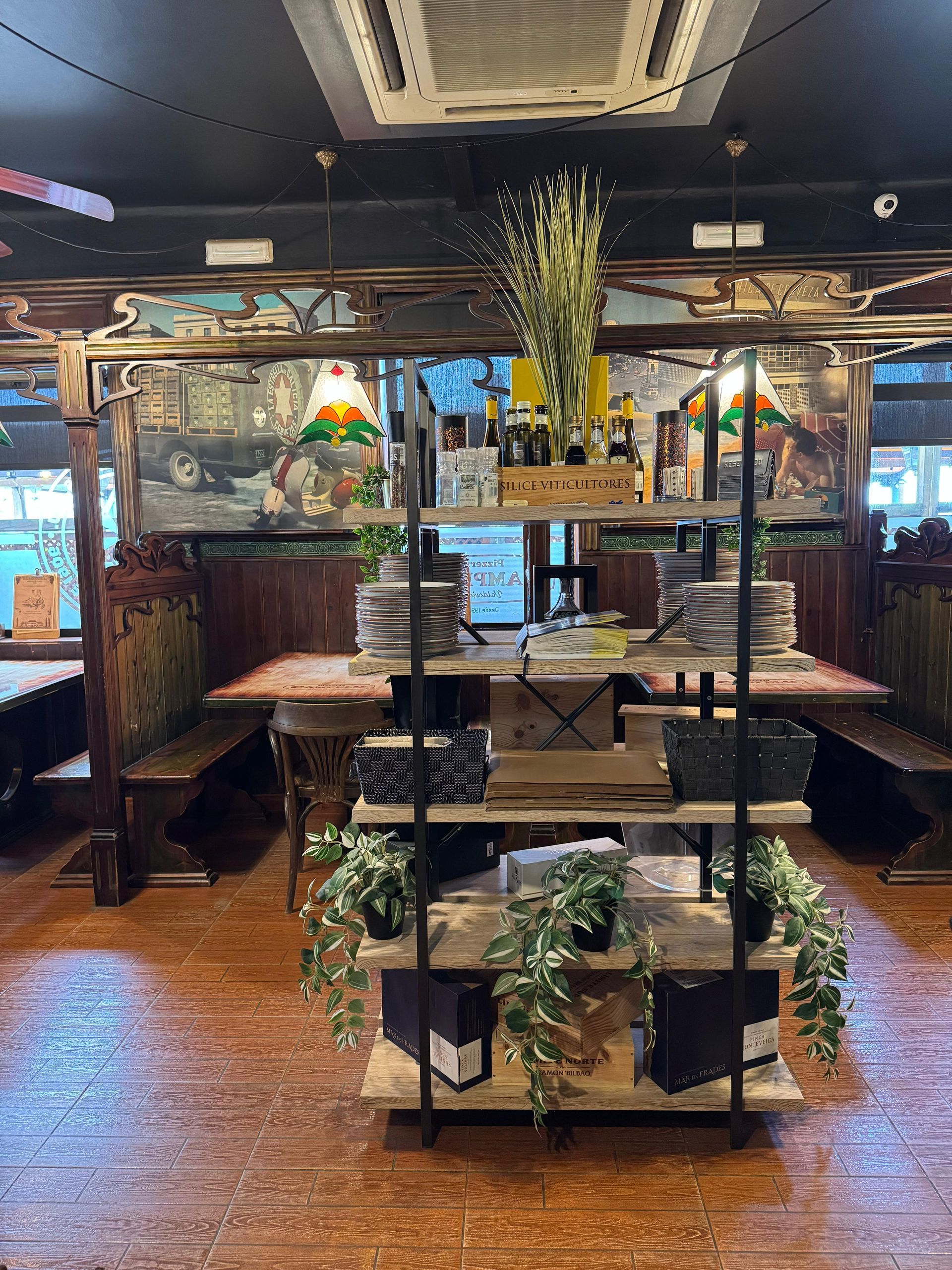 Interior de restaurante con mesas de madera, bancos y un estante de exhibición con plantas y productos.