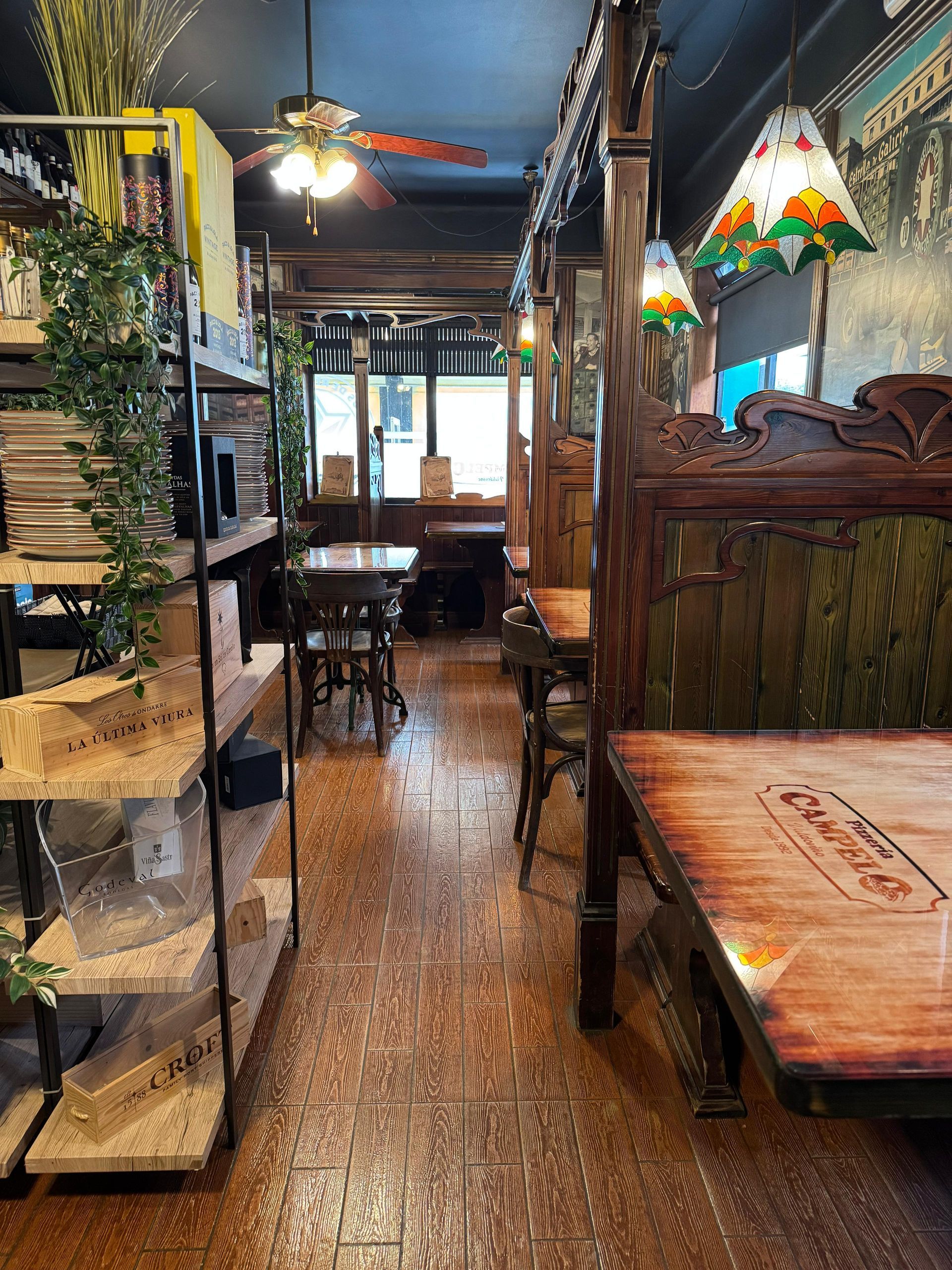 Interior del restaurante: mesas de madera, lámparas decorativas, estantes con artículos y un ventilador de techo parcialmente visible.