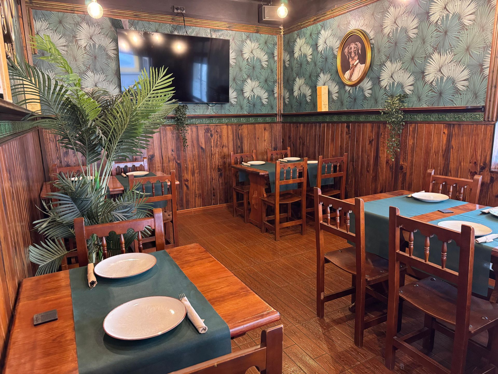 Interior de restaurante con paneles de madera, papel tapiz floral, mesas preparadas para cenar y un retrato en la pared.