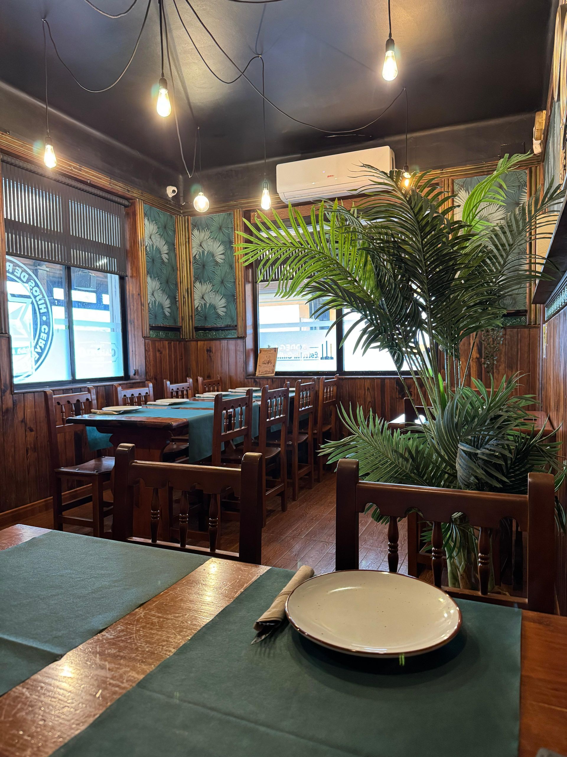 Interior de restaurante con mesas de madera, manteles verdes y plantas decorativas.