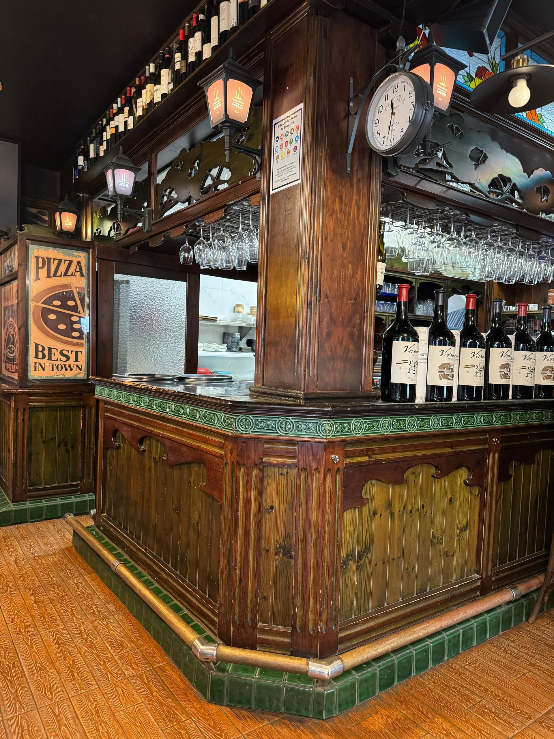 Barra de madera con botellas de vino, copas y un reloj. Tonos verdes y marrones; parece un restaurante.