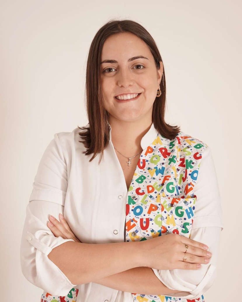 Una mujer con una bata blanca con letras está de pie con los brazos cruzados y sonriendo.