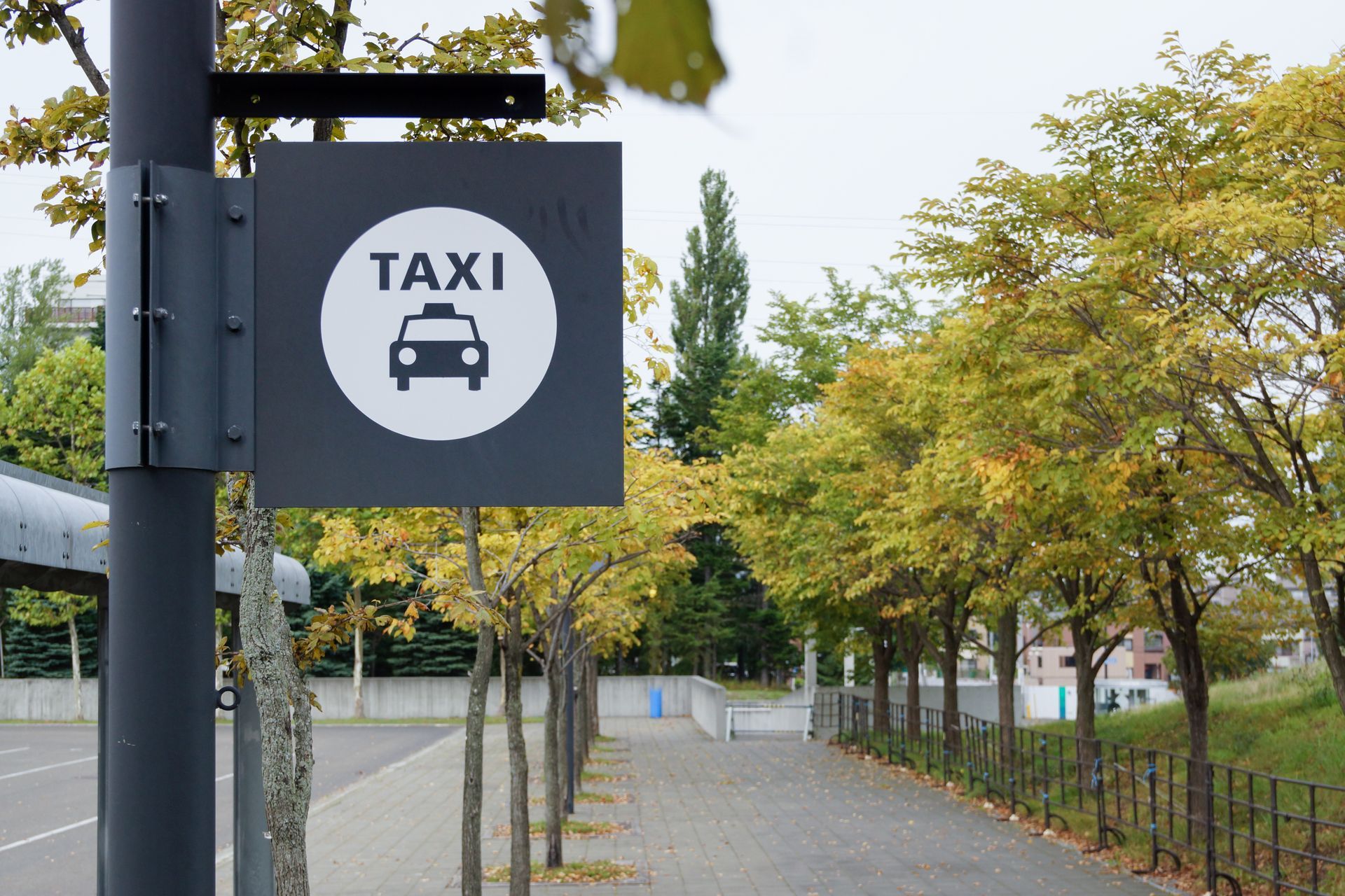 Un panneau de taxi carré gris avec un cercle blanc, une icône de taxi et du texte, fixé sur un poteau près d'un chemin bordé d'arbres.