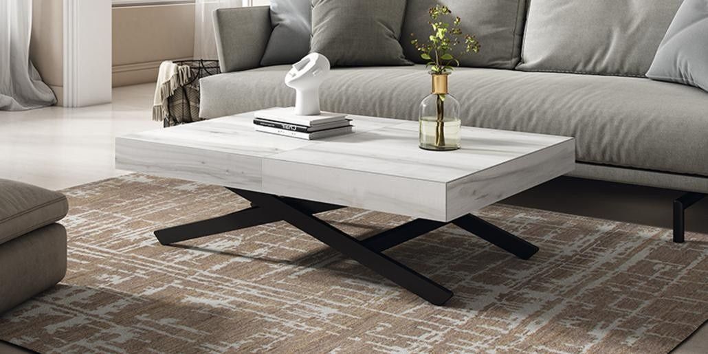 Mesa de centro elevable de color blanco con patas negras, sobre una alfombra estampada, frente a un sofá gris.