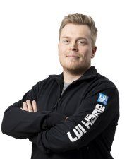 Hymyilevä henkilö mustassa vetoketjullisessa takissa kädet ristissä valkoista taustaa vasten