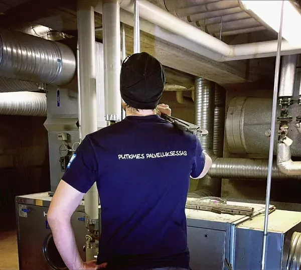 Tummansiniseen paitaan ja pipoon pukeutunut työntekijä tarkastaa teollisuuslaitteita mekaanisessa huoneessa, jossa on näkyvät kanavat.