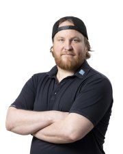 Mies mustassa poolopaidassa ja taaksepäin päin olevassa lippiksessä kädet ristissä valkoista taustaa vasten