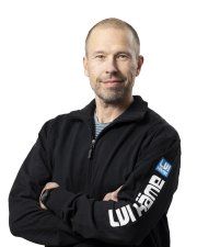 Mustaan takkiin pukeutunut mies seisoo kädet ristissä valkoista taustaa vasten.