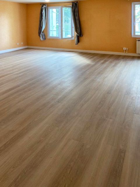 Pièce vide avec un sol imitation bois brun clair, des murs orange et deux fenêtres avec des rideaux gris.